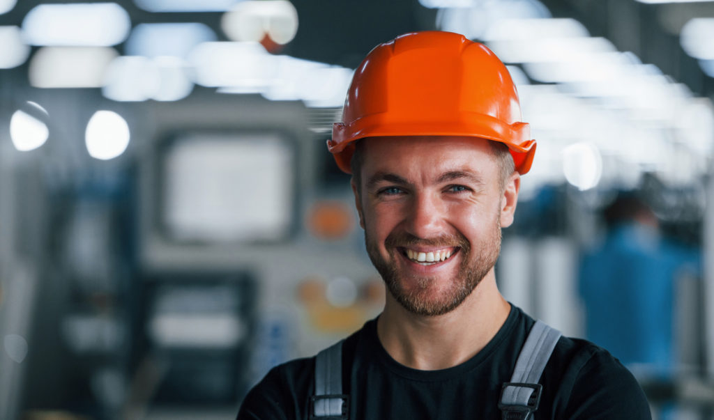 Smiling and happy employee. Portrait of industrial worker indoors in factory. Young technician with orange hard hat.