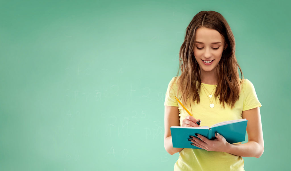 education, school, inspiration and people concept - smiling teenage student girl in yellow t-shirt writing to diary or notebook by pencil over green chalk board background