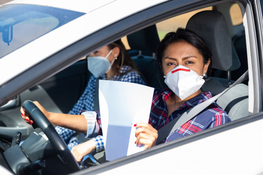 Portrait of middle aged Hispanic woman in medical face mask driving car, holding blank white sheet of paper, copyspace. Concept of control of coronavirus pandemic