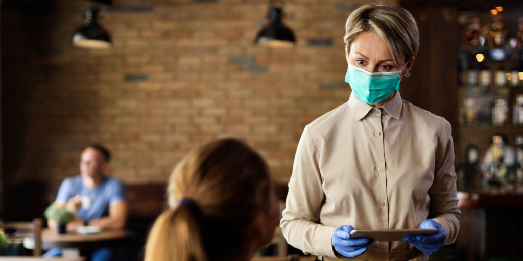 Waitress talking to a guest while using touchpad and wearing protective face mask due to COVID-19 pandemic.