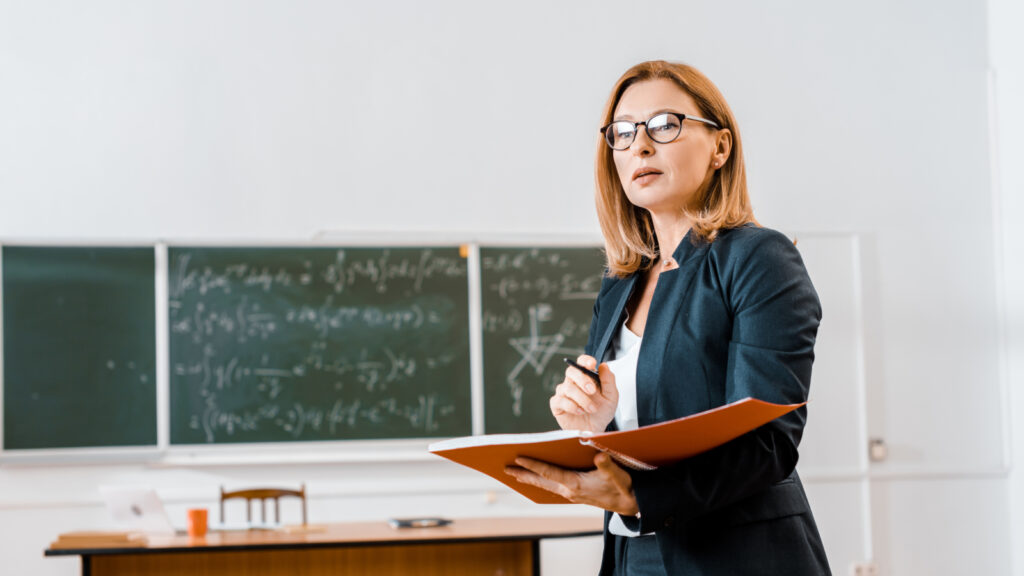 beautiful female teacher in formal wear and glasses holding notebook in classroom