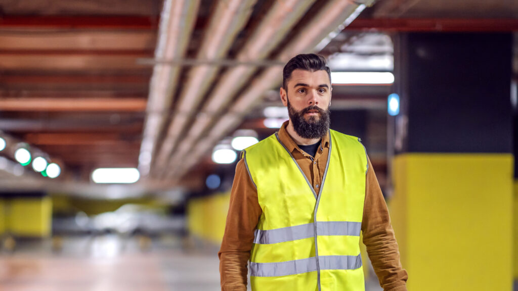 Three quarter length of handsome bearded worker in vest holding his safety helmet and going back to his workplace. Underground garage in construction process interior.
