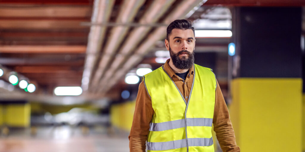 Three quarter length of handsome bearded worker in vest holding his safety helmet and going back to his workplace. Underground garage in construction process interior.