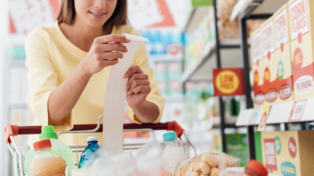 Smiling woman doing grocery shopping at the supermarket and checking a long receipt, she is leaning on the full cart